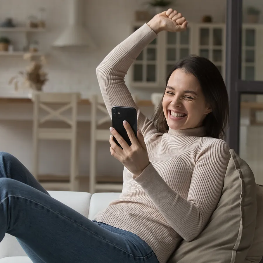 Junge Frau freut sich zu Hause über eine Nachricht auf ihrem Smartphone und nutzt dabei die Jobcenter App.